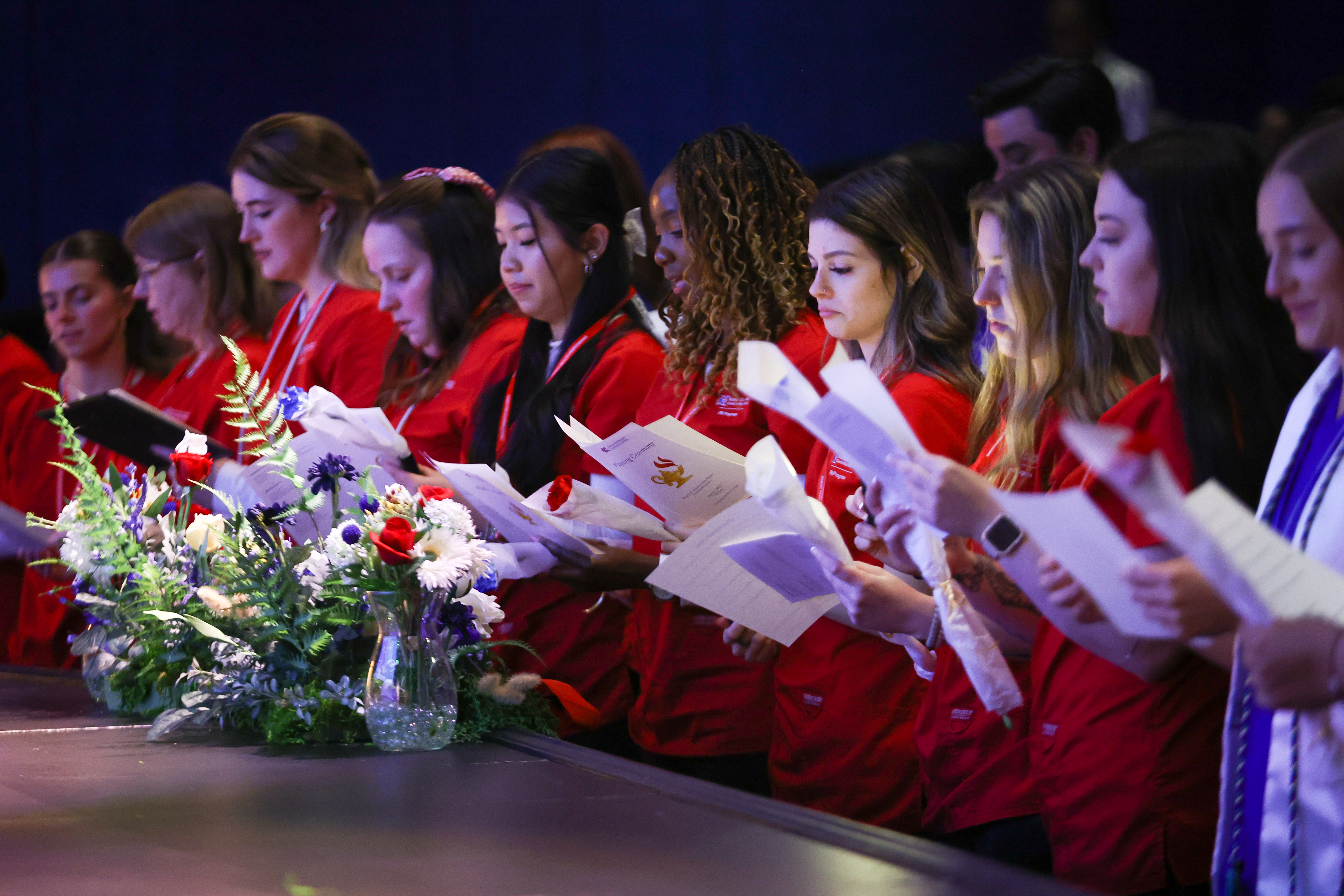 Students in red scrub tops in a line reading from program.